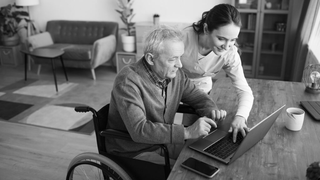 En äldre man sitter i rullstol vid ett bord i ett vardagsrum och knappar på en laptop. En kvinna hjälper honom att navigera.